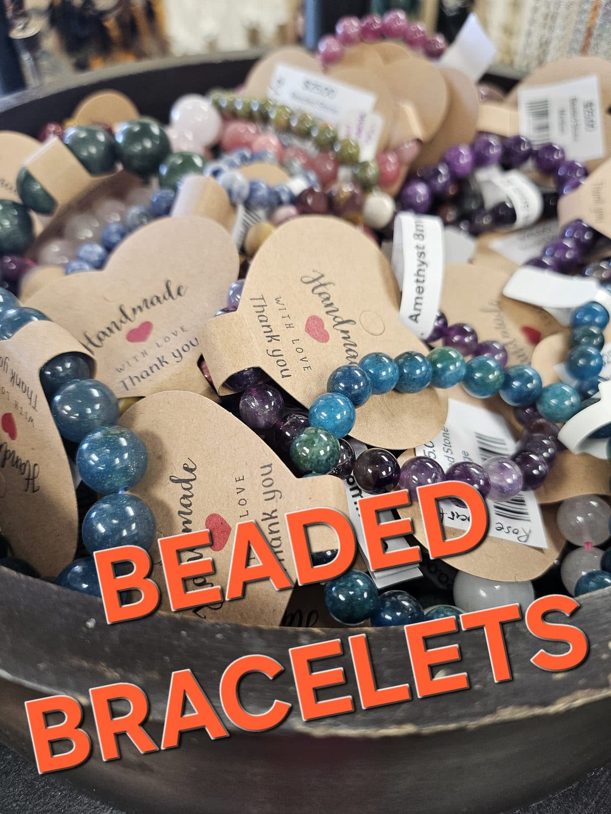 A wooden bowl heaped with gemstone bracelets on kraft “Handmade with love” tags, captioned “Beaded Bracelets”.