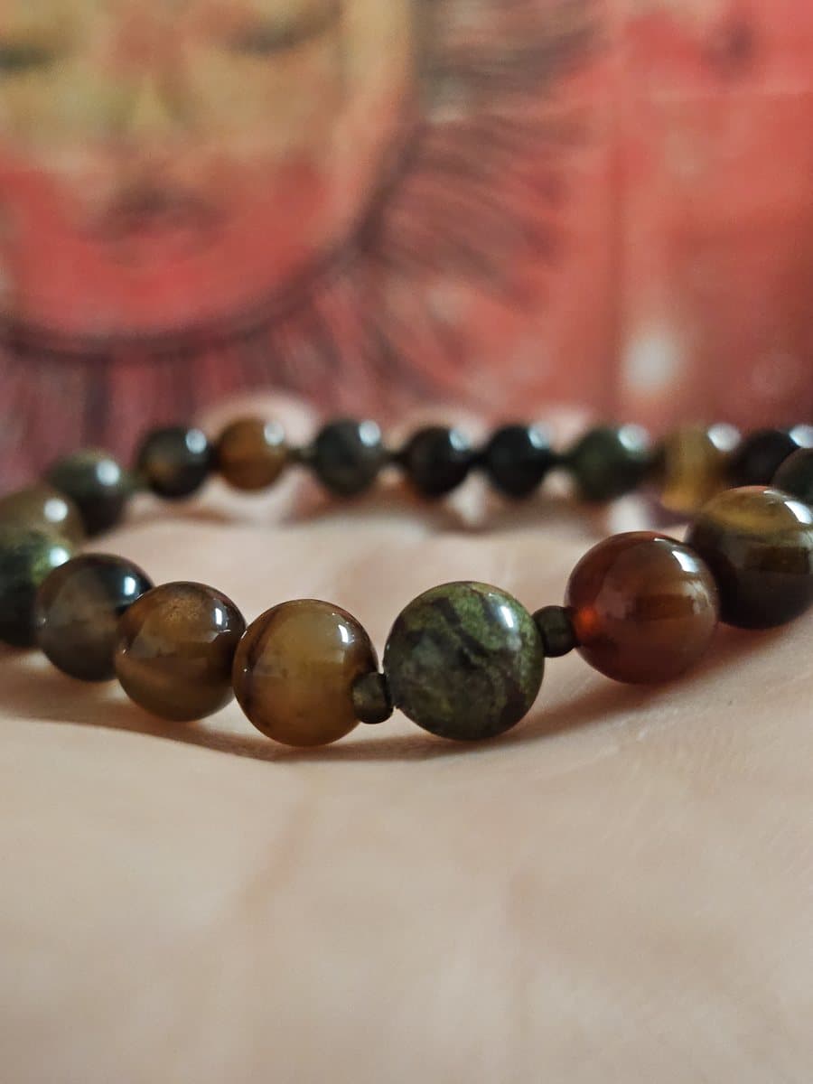 A beaded bracelet of polished green and amber stones resting on an open palm, with a painted sun behind it.