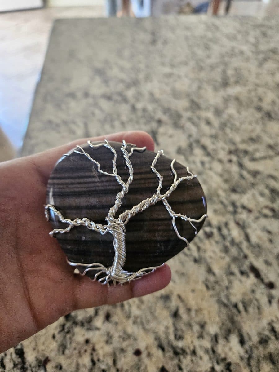A silver wire tree of life wrapped across a banded grey stone, held in the palm of a hand.